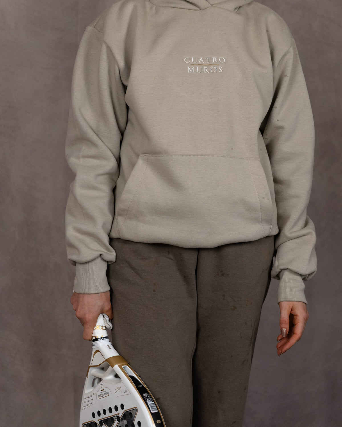 The Malla Hoodie in Natural Stone, front with a padel racket, worn by a 5ft 6 model in size Small