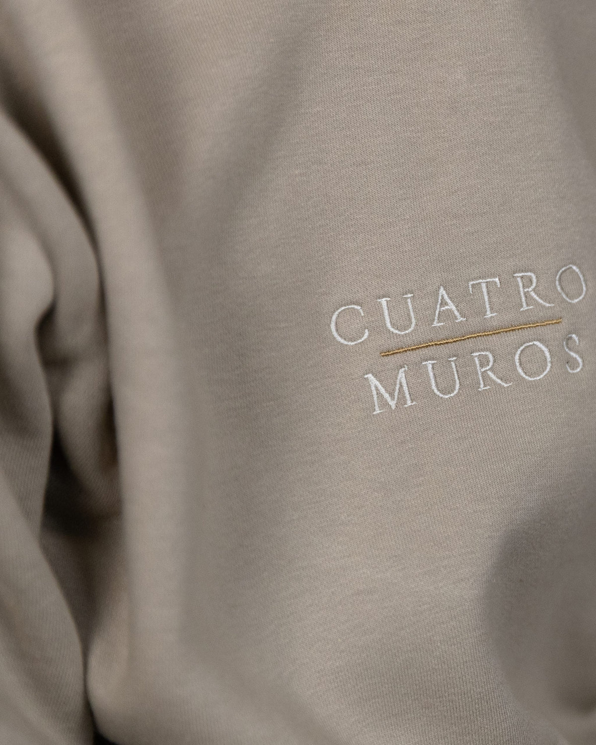 The Malla Hoodie in Natural Stone, close-up of the stacked wordmark embroidered in Oat Milk with a Desert Sand rule