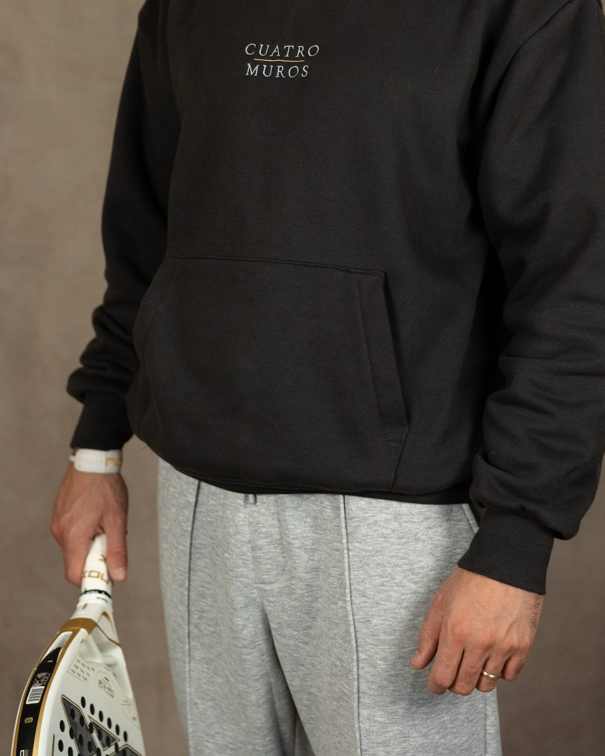 The Malla Hoodie in Solid Charcoal, front with a padel racket, worn by a 6ft model in size Large