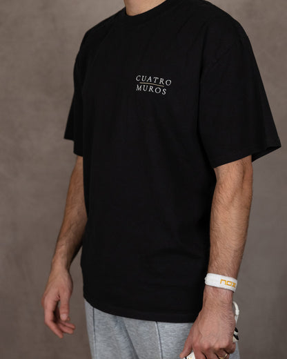 The Muro Tee in Faded Black, three-quarter front with a padel racket, worn by a 6ft model in size Large