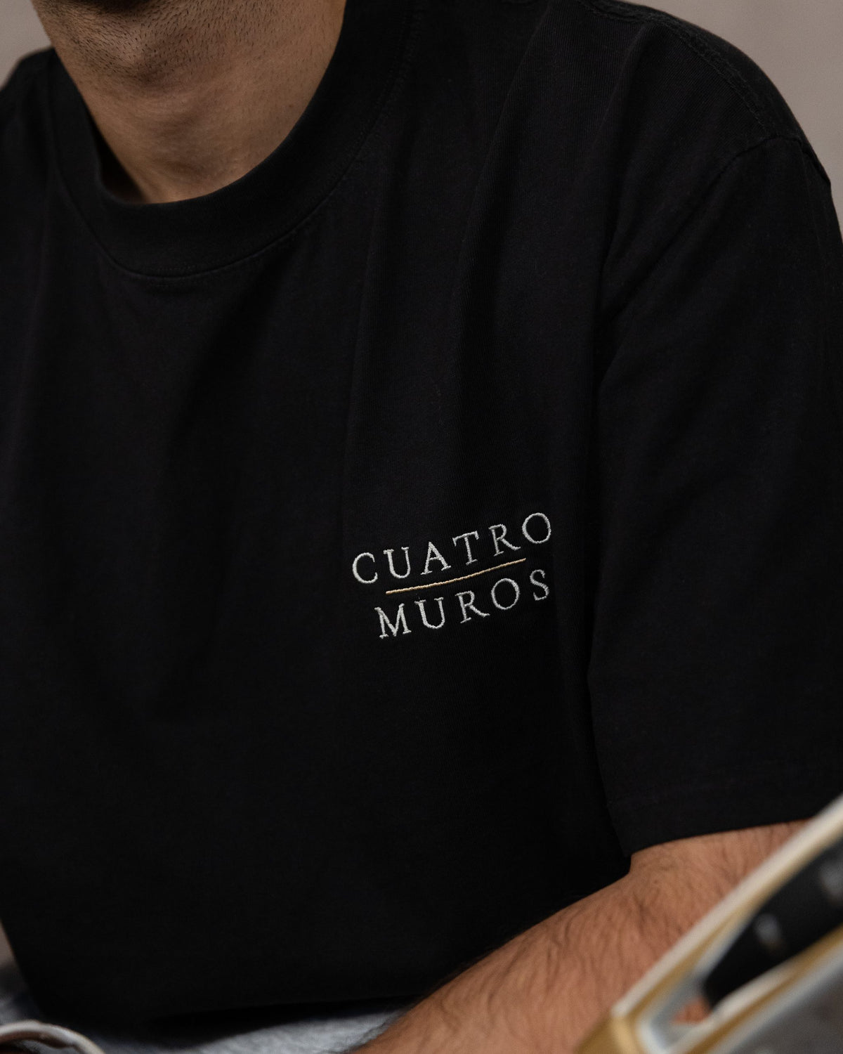 The Muro Tee in Faded Black, close-up of the stacked wordmark embroidered in Oat Milk with a Desert Sand rule