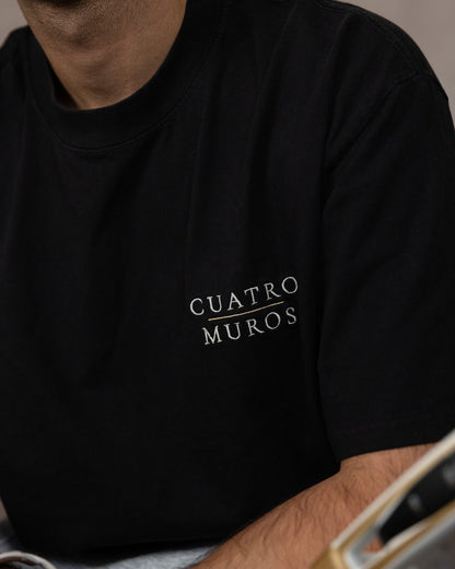 The Muro Tee in Faded Black, close-up of the stacked wordmark embroidered in Oat Milk with a Desert Sand rule